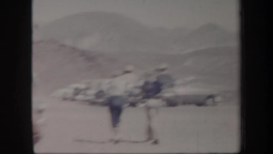 Soft 8mm view of visitors and cars in an Arizona outdoor parking area, with pale hills in the distance.