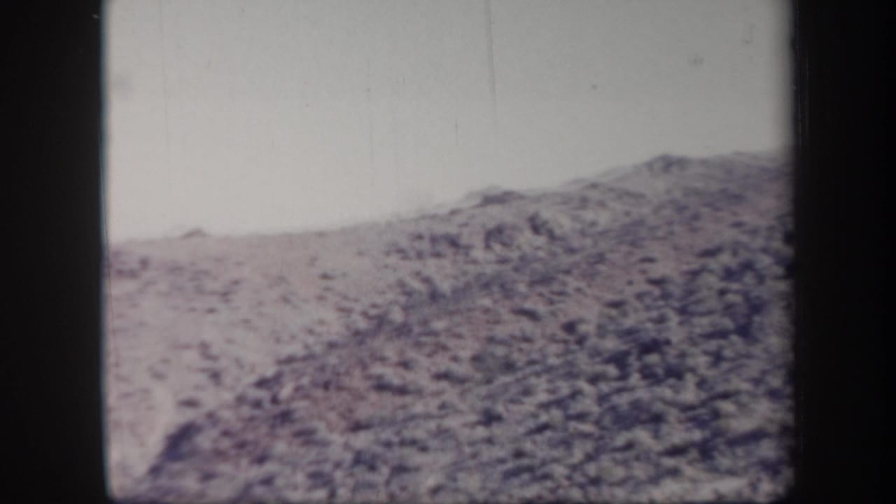 1957 Arizona 8mm frame showing a dry rocky hillside under a pale sky, with soft focus and faded color.