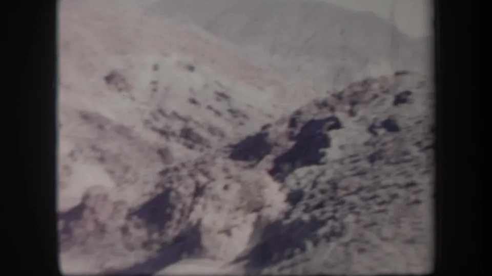 1957 Arizona home-movie frame showing a dry hillside, dark rock formations, and visible 8mm grain.