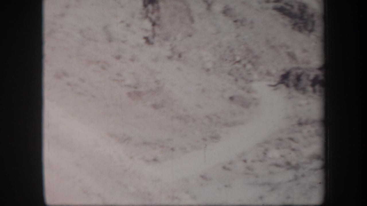1957 Arizona 8mm frame showing a pale sandy desert slope and rocky hillside, with no person clearly visible in this frame.