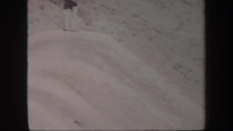1957 Arizona 8mm frame of a pale wind-shaped sand dune below a rocky desert slope, with no distinct person visible.