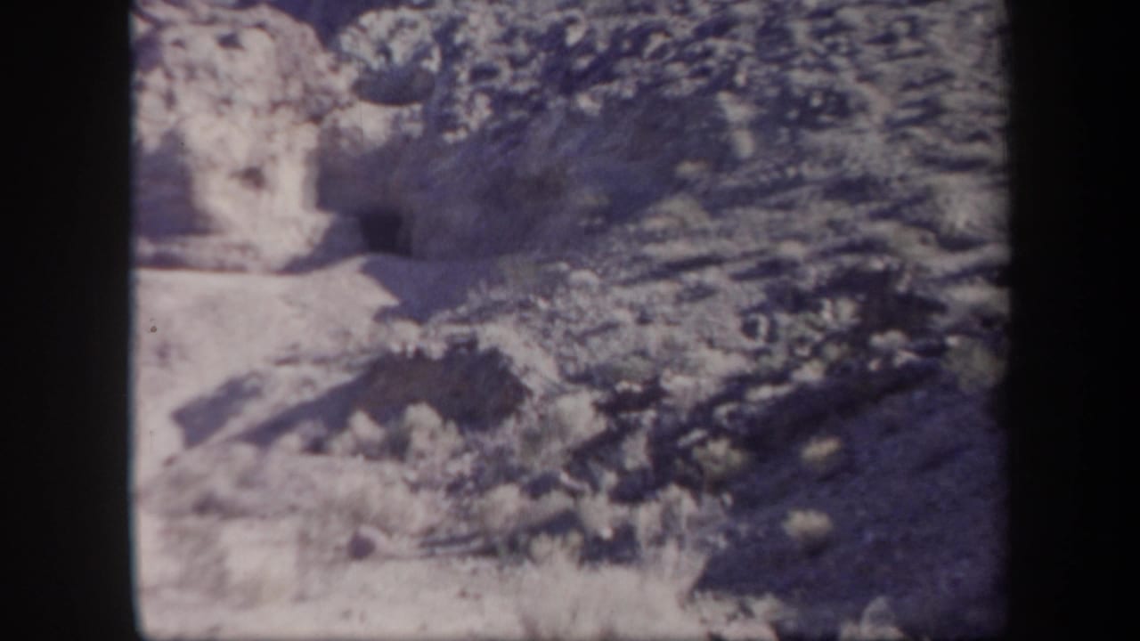 1957 Arizona 8mm frame showing arid rocky terrain, scrub growth, and a dark cave-like opening set into a cliff face.