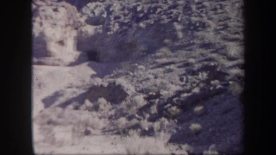 1957 Arizona 8mm frame showing arid rocky terrain, scrub growth, and a dark cave-like opening set into a cliff face.