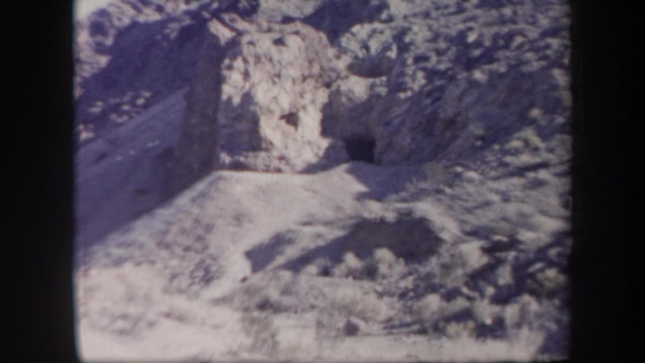 1957 Arizona home-movie frame with a dark cave-like entrance in a rocky cliff above pale desert ground.