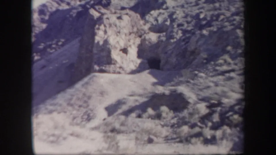 1957 Arizona home-movie frame with a dark cave-like entrance in a rocky cliff above pale desert ground.