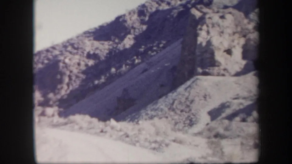 1957 Arizona 8mm frame showing a rugged arid slope, pale rocky ground, and sparse scrub below a cliff face.