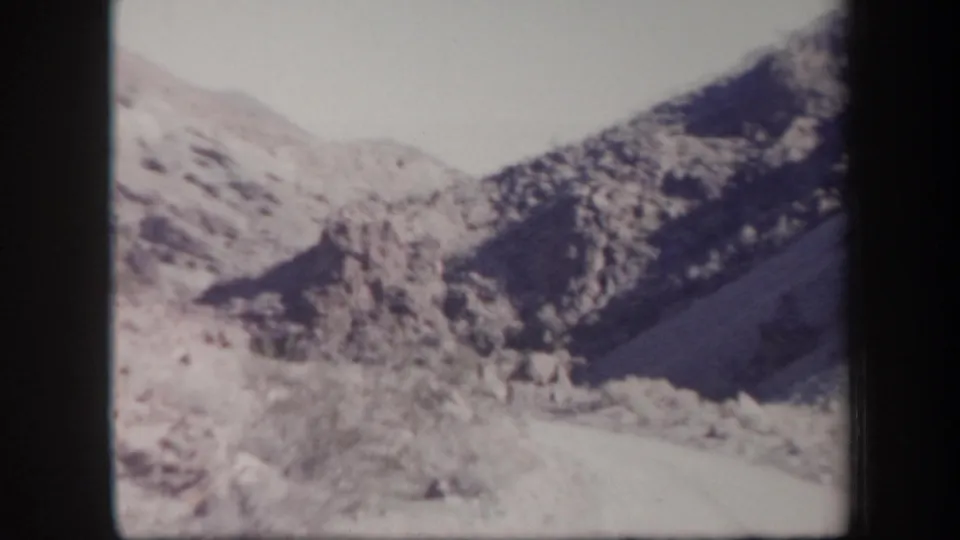 1957 Arizona home-movie frame with distant rocky ridges, scrub-covered slopes, and a pale road or wash in the foreground.