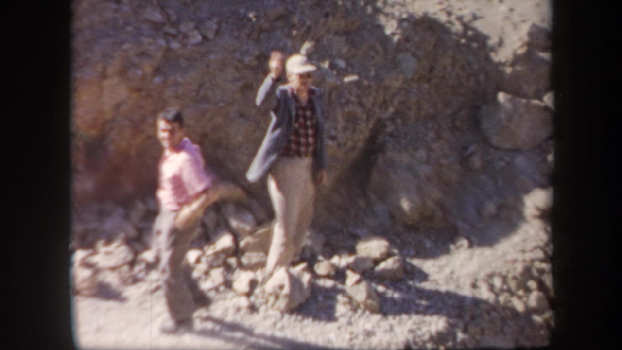 Two adult men on a rocky Arizona hillside, one in a hat and jacket waving toward the 1957 home-movie camera.
