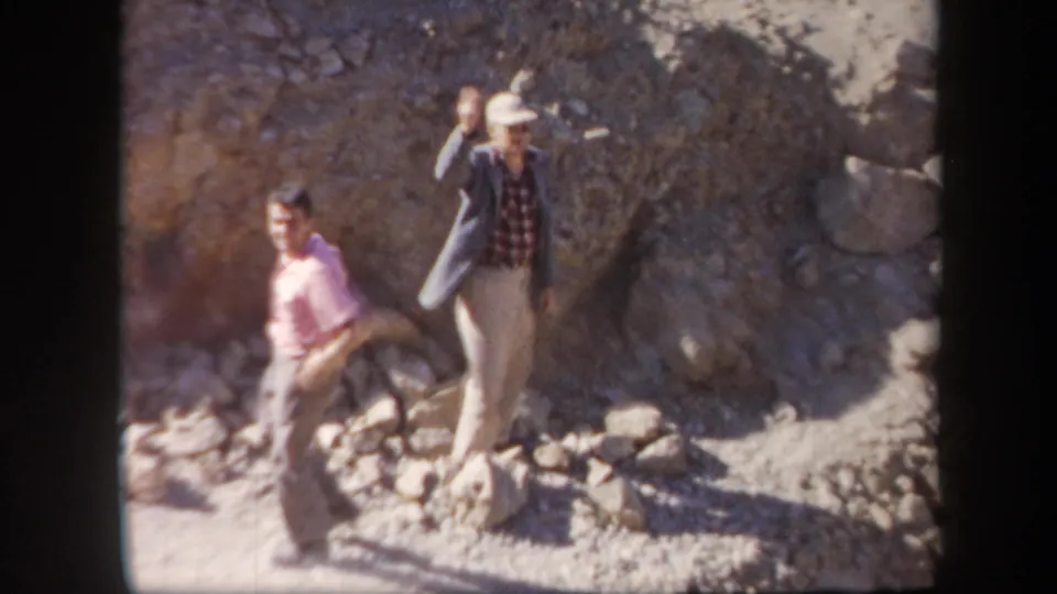 Two adult men on a rocky Arizona hillside, one in a hat and jacket waving toward the 1957 home-movie camera.