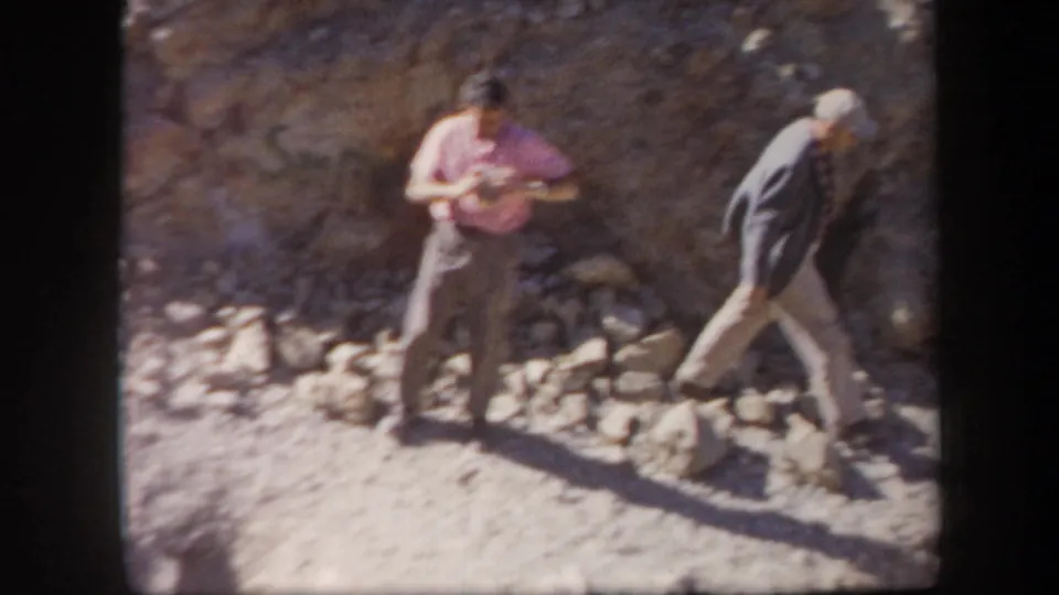 Two adult men in casual mid-century clothing move carefully across loose rocks on an Arizona hillside.