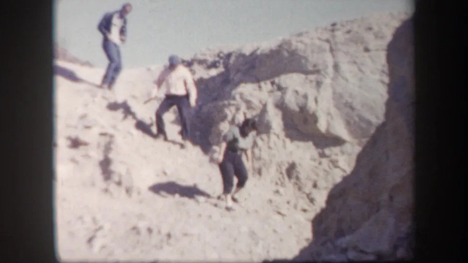 Several adult men descend a steep sunlit rocky hillside in 1957 Arizona home-movie footage.