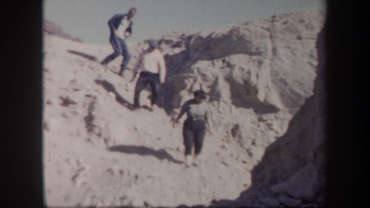 1957 Arizona home-movie frame showing three adults moving carefully down a sunlit sandy desert slope.
