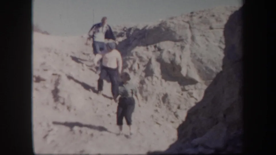 The group continues descending a rugged Arizona desert incline, with pale rock and strong shadows visible.