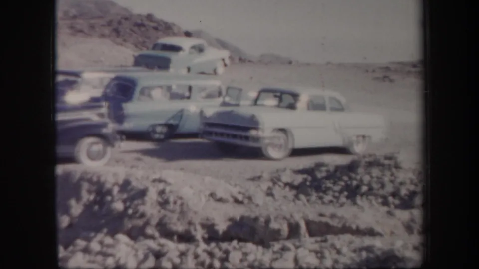 Several mid-century cars, including a light sedan, sit near a rocky desert roadside in Arizona home-movie footage.