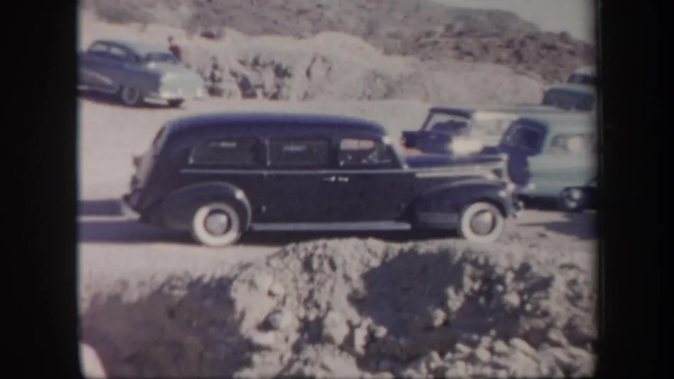 A dark mid-century station wagon is seen beside other cars and rocky Arizona desert terrain in faded 8mm footage.