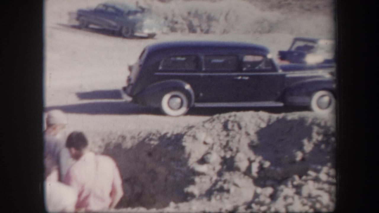 1957 Arizona home-movie frame showing a dark station wagon on a dirt road near rocks, with tourists partly visible in bright sun.