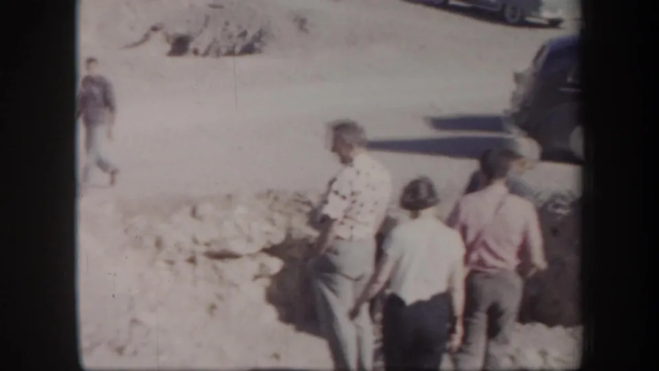 1957 Arizona frame with several tourists walking through a sunlit rocky area near parked cars at a scenic or historic site.