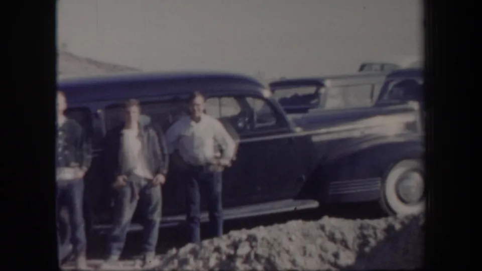 1957 Arizona home-movie frame with men gathered beside a dark station wagon at a rocky roadside location.