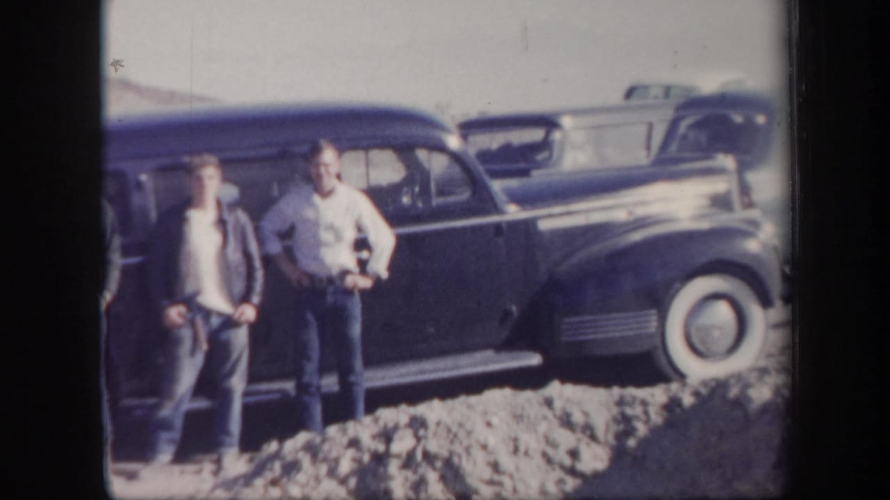 Two young men stand near a dark vintage sedan in a desert setting.