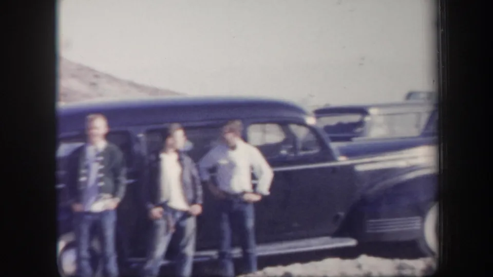 Three young men stand in front of a dark sedan in a 1957 Arizona desert scene, with another vintage car visible in the background.