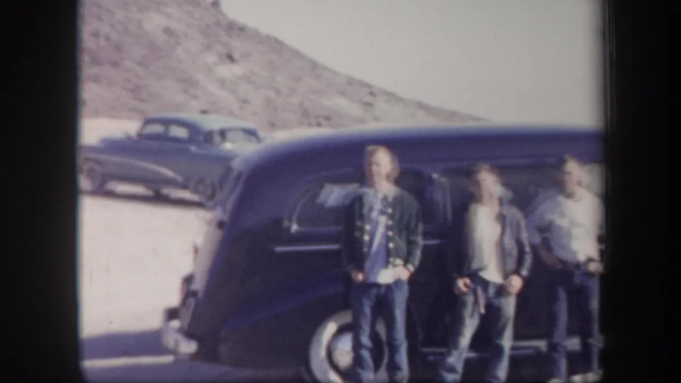 Three young men are framed beside a dark vintage sedan in the Arizona desert, with another car and low hills behind them.