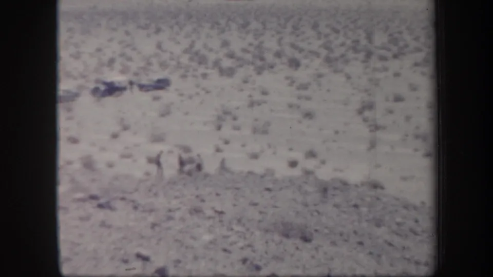 A soft 1957 Arizona desert view shows scrubby ground and hillside, with a small light-toned object or figure near the lower center.