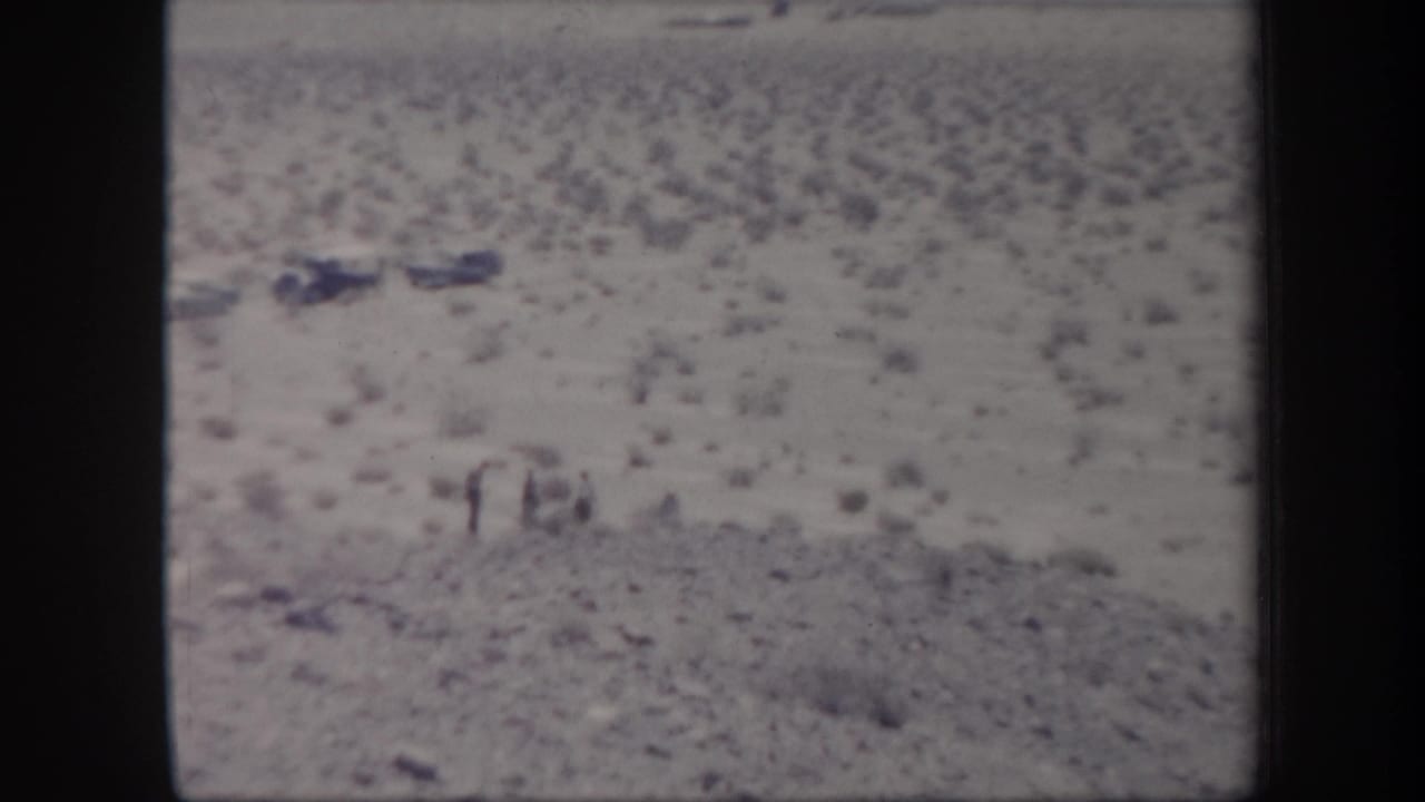 1957 Arizona 8mm frame showing small figures in open desert terrain near several parked mid-century cars.