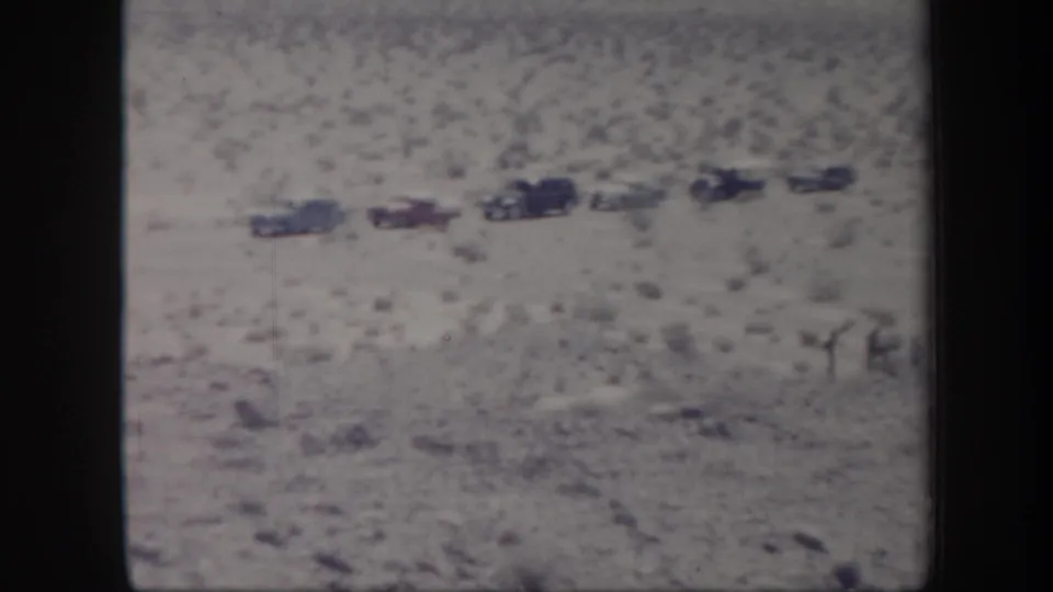 Soft 8mm frame of a 1957 Arizona desert stop, with several classic cars and small figures in open brushland.