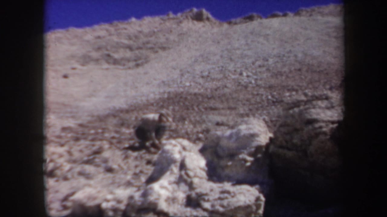 1957 Arizona 8mm frame showing a person crouched near white rocks on a dry rocky hillside beneath a bright blue sky.