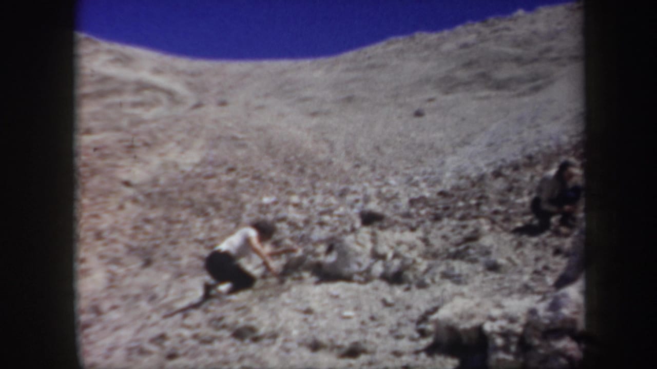 Person kneeling on a rocky slope with another figure partly visible at the edge.