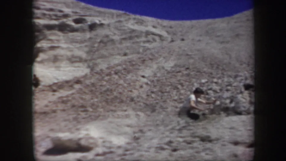 Wide 8mm view of a person crouched on a white rocky Arizona hillside with dry terrain rising behind them.