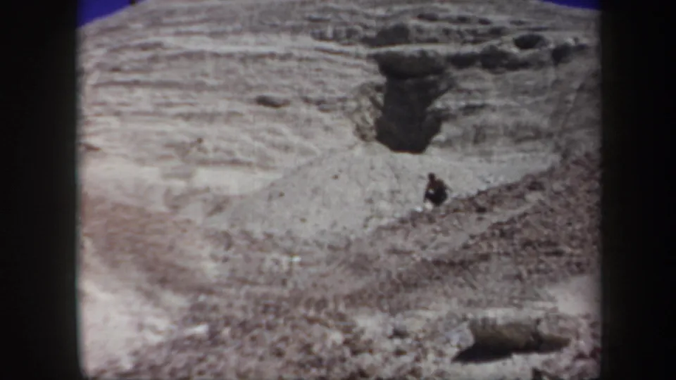 A small figure stands or crouches on a pale eroded Arizona hillside below a dark opening or shadowed recess in the rock.