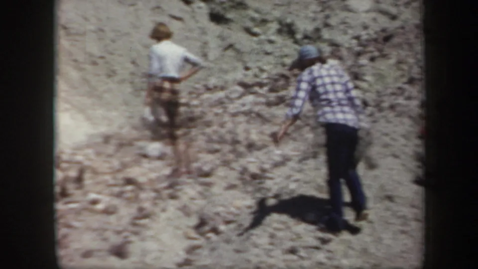 Arizona 1957 home-movie frame showing two family members in hats walking up a rough, sunlit mountain slope.