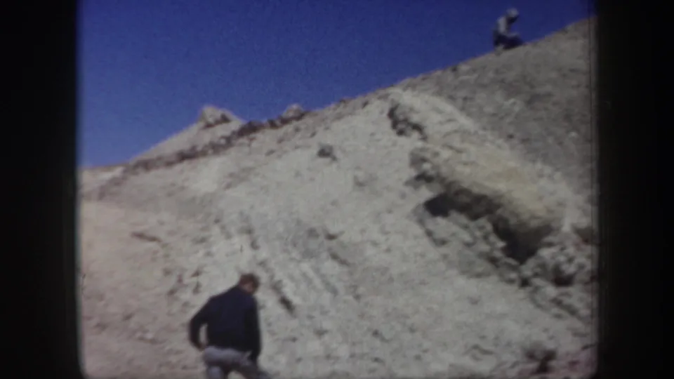 Arizona 1957 home-movie frame with one person on a pale rocky slope and another small figure near the ridgeline under blue sky.