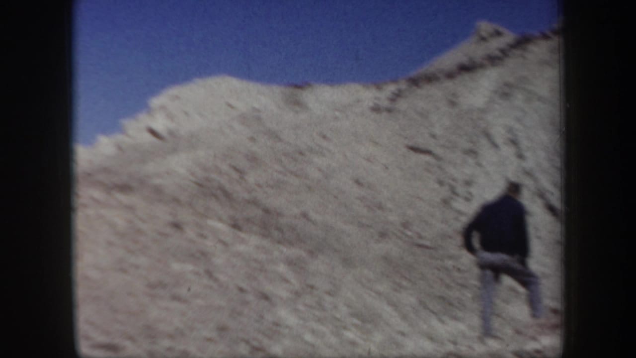1957 Arizona 8mm frame showing an adult man crossing a light-colored rocky desert slope under clear sky.