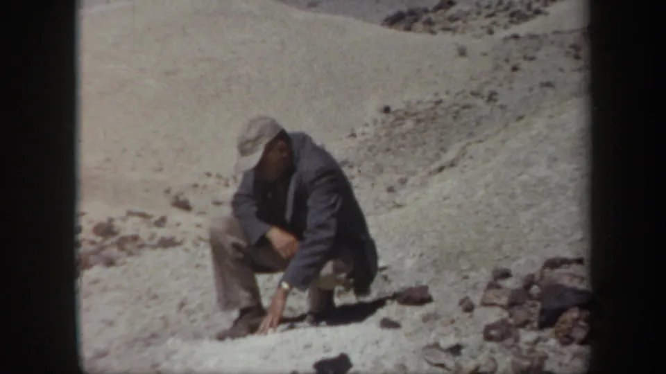 1957 Arizona 8mm frame showing an adult man crouched on a desert slope to examine stones on the ground.