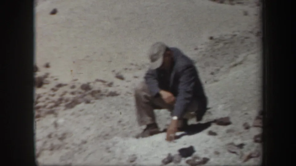 1957 Arizona 8mm frame showing an adult man kneeling or crouching near rocks in a sunlit desert setting.