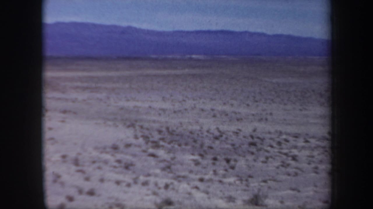1957 Arizona 8mm frame looking across a flat, barren desert plain toward distant blue mountain ridges under a clear sky.