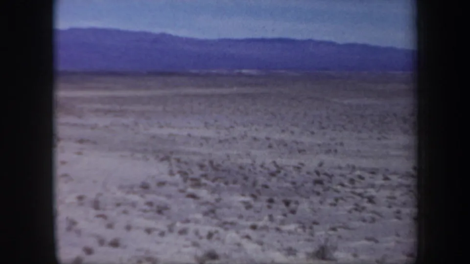 1957 Arizona 8mm frame looking across a flat, barren desert plain toward distant blue mountain ridges under a clear sky.