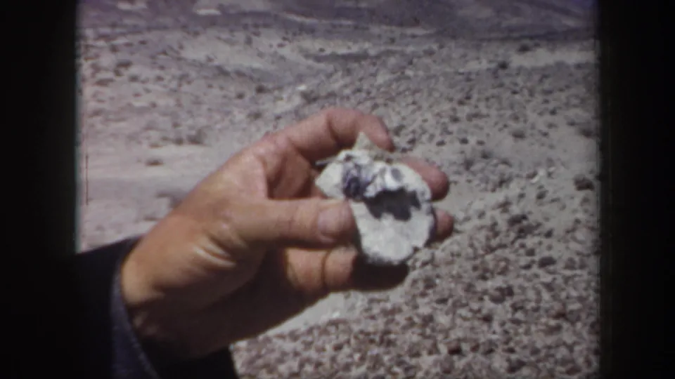 1957 Arizona 8mm frame showing an adult hand holding a light-colored rock specimen against rocky desert terrain.