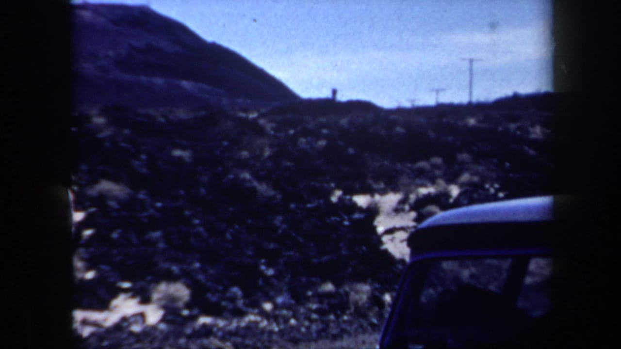 Dark rocky desert terrain with a low hill, utility poles, and part of a car.