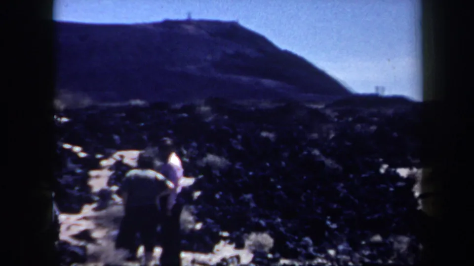 A small group of adult visitors walks through rocky Arizona desert terrain below a low hill, filmed from a handheld 8mm travel perspective.