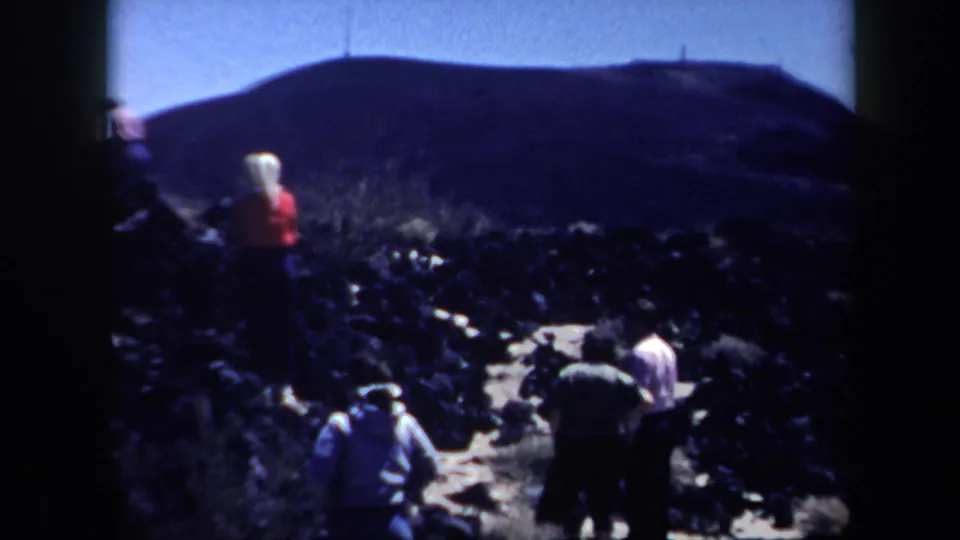 Adult visitors in casual clothing explore a rocky Arizona desert path, with scrub, dark stone, and a low hill visible in the background.