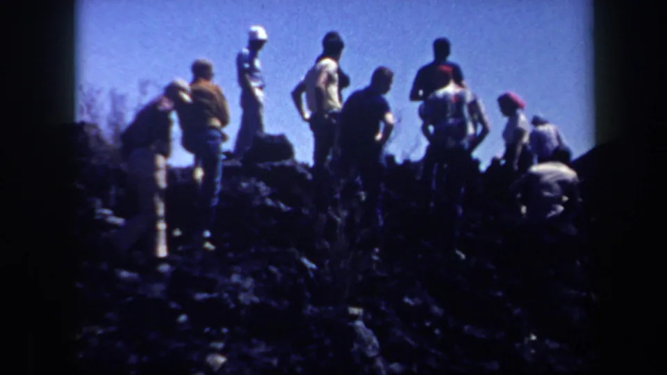 1957 Arizona footage frame with several adults standing or crouching on a rough hillside, filmed from a low handheld angle.