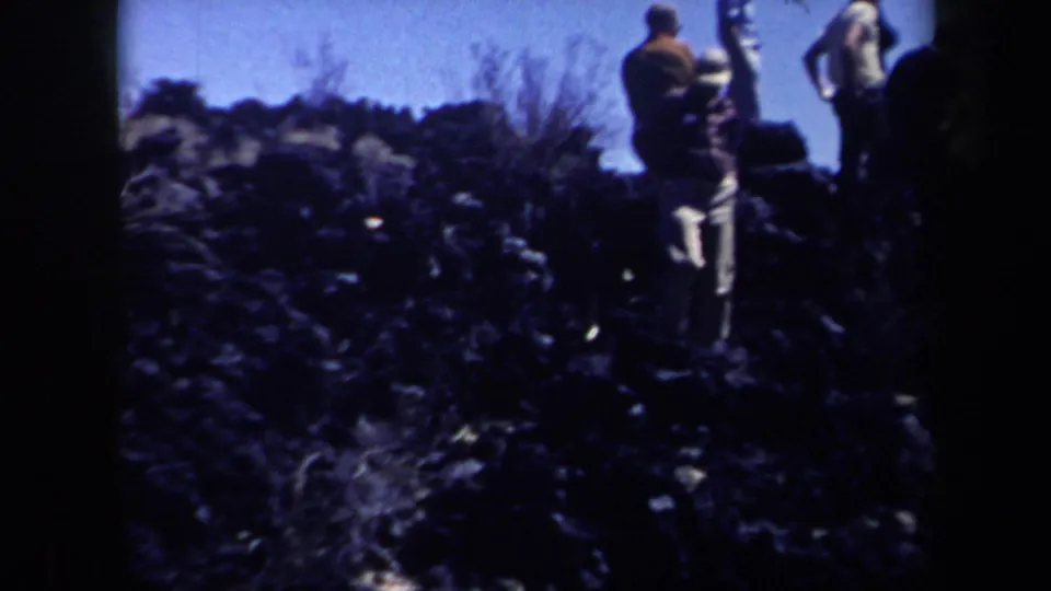 A dark, grainy 8mm frame shows people moving through rocky Arizona terrain with brush, strong shadows, and a bright sky.