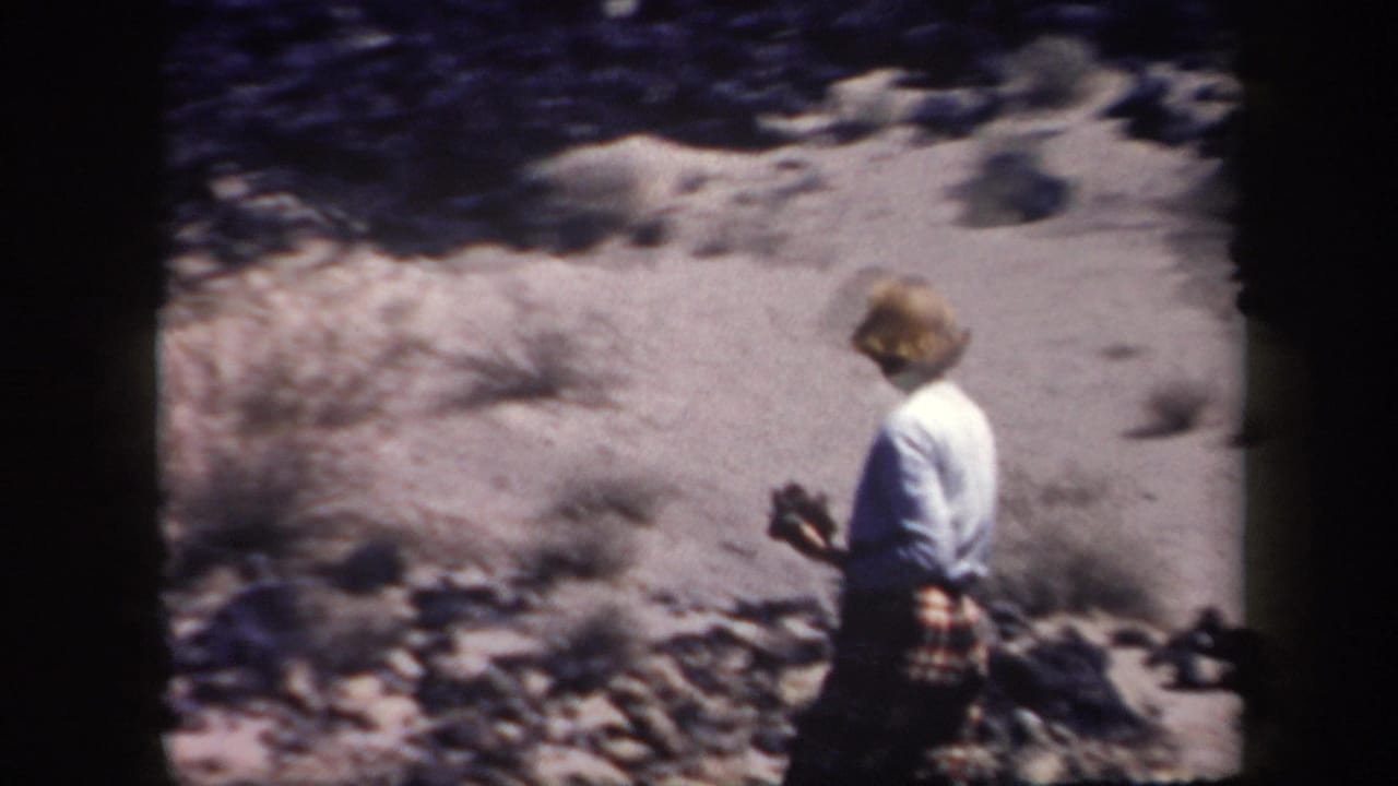 1957 Arizona home-movie frame of a woman walking through sparse rocky desert terrain in a light shirt and plaid skirt.