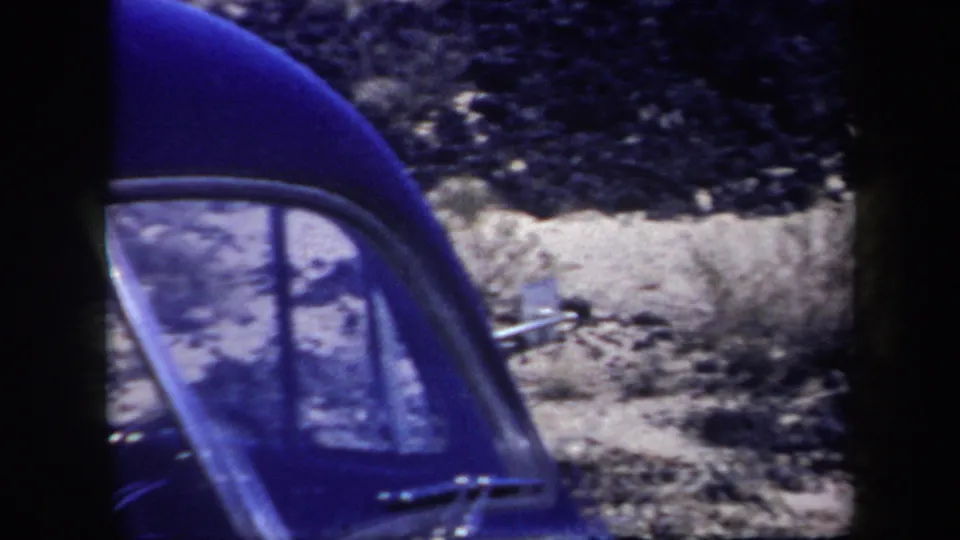 Faded 1957 Arizona 8mm frame with a partial car window at left and rocky desert ground beyond it.