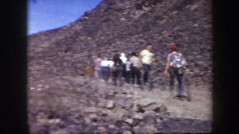 A group of adult men walks away along a rocky Arizona mountain trail in grainy 1957 home-movie footage.