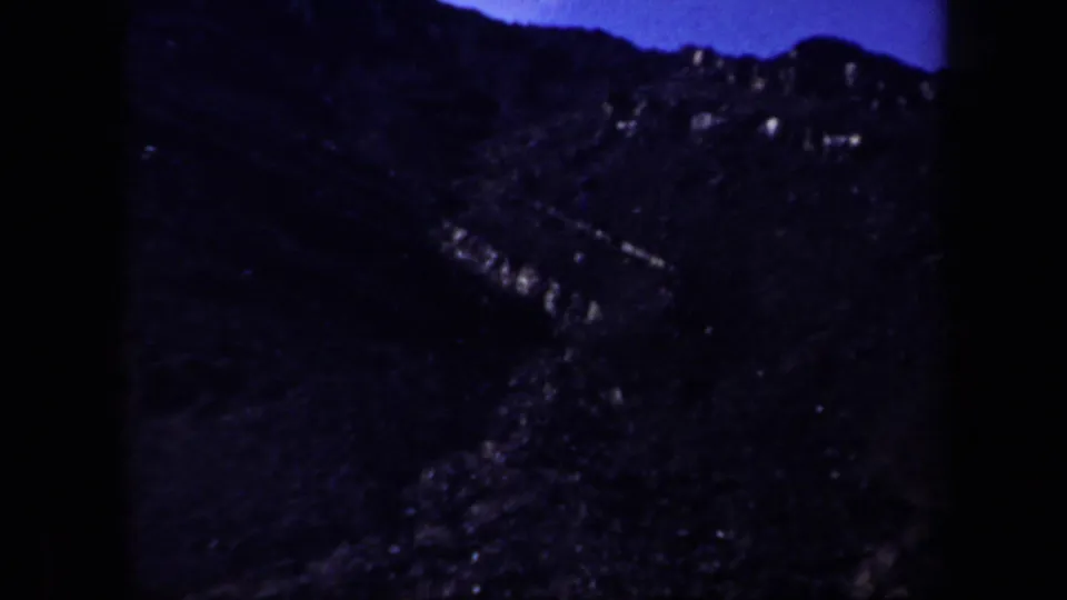 1957 Arizona 8mm frame showing a dark, rugged hillside with pale rock streaks under a deep blue sky.