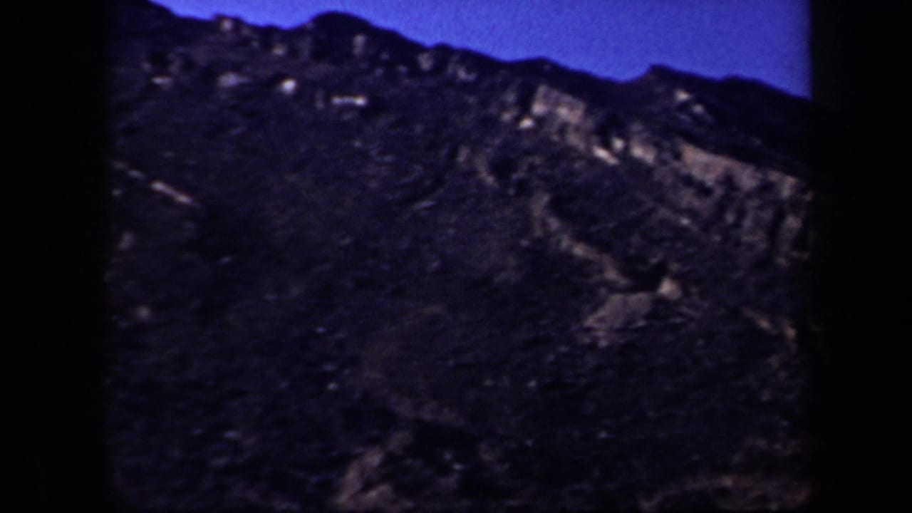 Rocky mountain slope with layered outcrops and a dark ridgeline against blue sky.